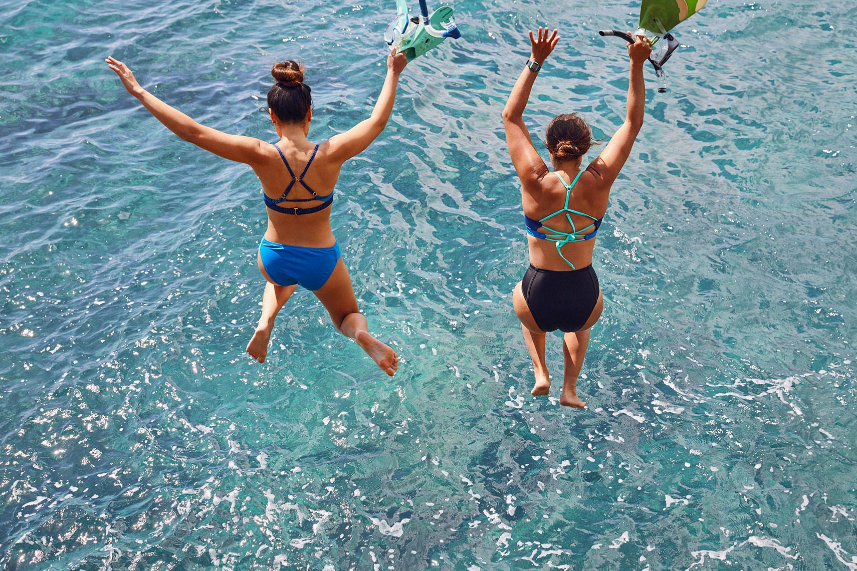 two women jumping into ocean in swimwear with hands overhead