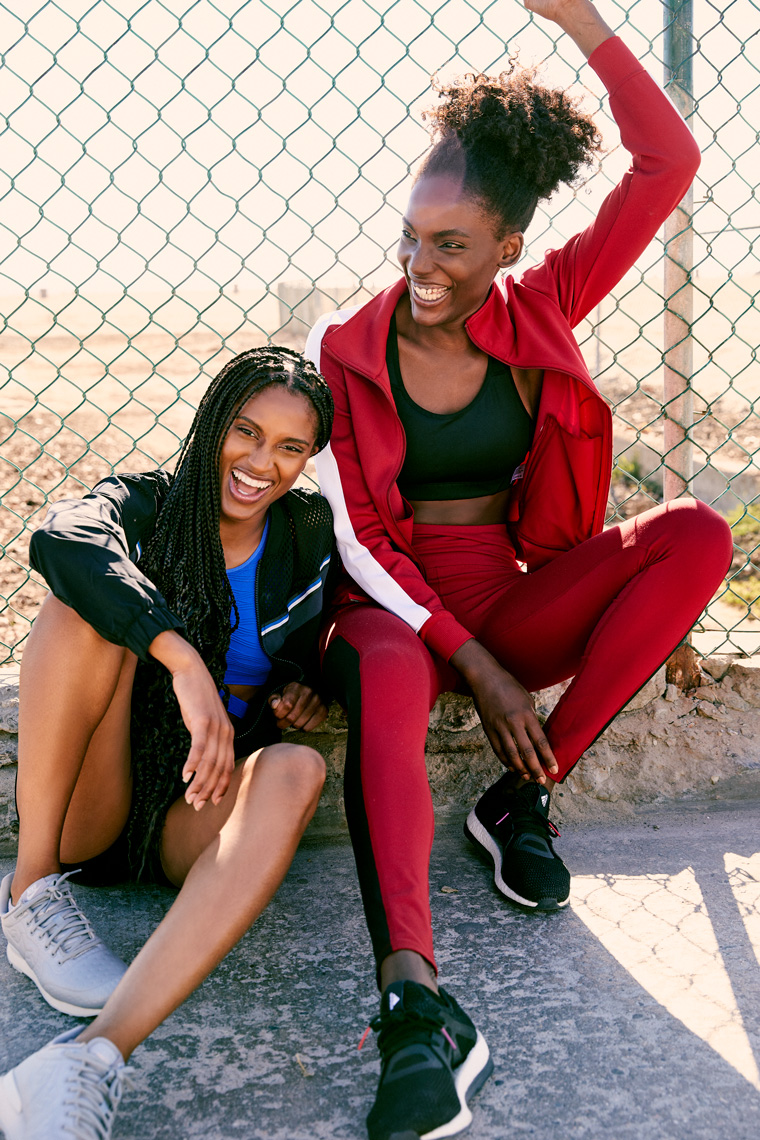 two women in athletic wear sitting against a fence and laughing 